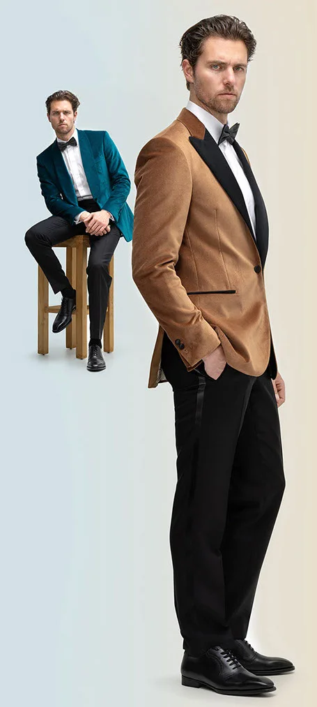 Two male models in two-piece suits with black bow ties; one standing in brown blazer and black pants, the other seated in dark green blazer and black pants on a wooden bench.