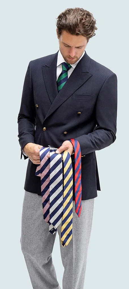 Male model in black blazer and light pants holding four ties with navy diagonal stripes in pink, white, yellow, and red.