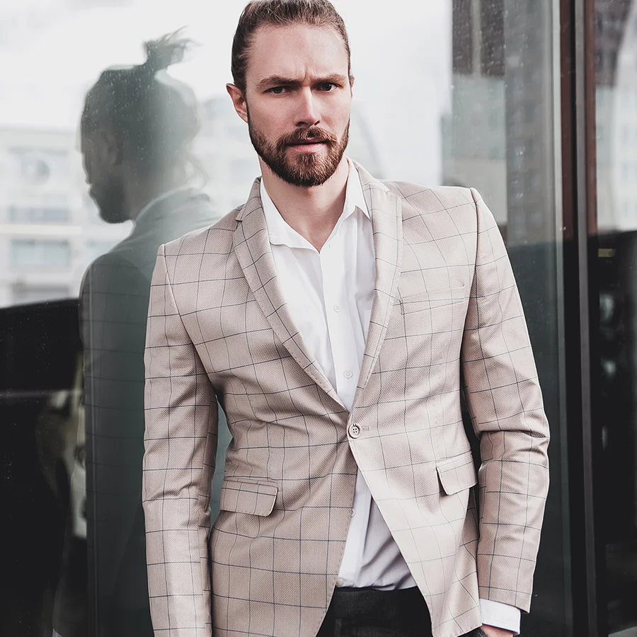 Male model wearing beige checked blazer and white shirt with low bun hairstyle, standing confidently with glass reflection behind him.