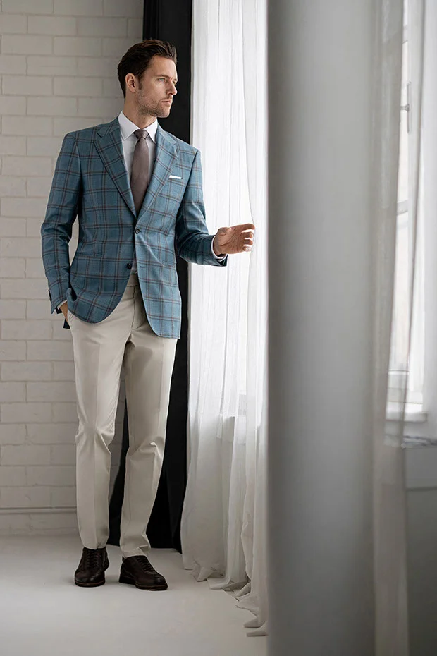 Model pulling back curtains wearing blue windowpane suit jacket, beige pants, brown shoes, light shirt, and brown tie.