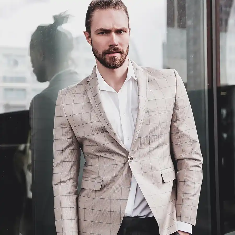 Male model wearing beige checked blazer and white shirt with low bun hairstyle, standing confidently with glass reflection behind him.
